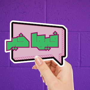 May include: A sticker with Arabic text in green, outlined in purple, inside a pink speech bubble. The speech bubble has a black border and is held against a purple brick wall. The sticker is held by a hand with a purple background.