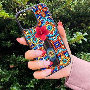 May include: A black phone case with a colorful stained-glass design. The case features a red flower and a graphic of a bottle with the text "Coffee Solace". The case is held against a backdrop of green foliage.