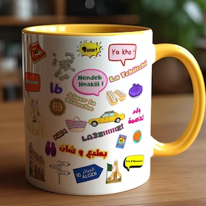 Könnte beinhalten: Weißer Keramikbecher mit gelbem Innenraum und Henkel, verziert mit bunten Aufklebern und Text. Die Aufkleber enthalten Sätze wie "Ya kho!" und "Alger la Blanche" sowie verschiedene Illustrationen. Der Becher steht auf einer Holzoberfläche.