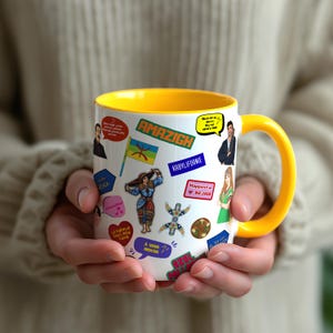 May include: A white ceramic mug with a yellow interior and handle, decorated with colorful stickers and text. The stickers feature illustrations, flags, and phrases, including "AMAZIGH" and "Happiest in Bejaia". The mug is held by a person.