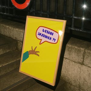 May include: A framed yellow artwork with a speech bubble containing text. The speech bubble is pink with blue text. A cartoon hand with a green sleeve points towards the text. The artwork is framed in light wood.