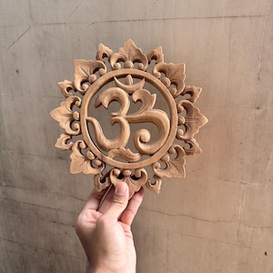 Op de afbeelding: Een houten wanddecoratie met een gegraveerd Om-symbool in het midden, omringd door een decoratief bloemmotief.