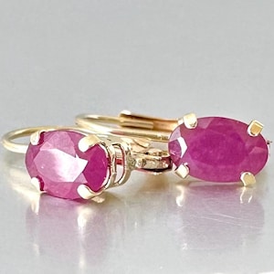 以下が含まれることがあります： オーバルカットのルビーをあしらった、ゴールドのレバーバックピアス。ルビーは鮮やかなピンク色で、磨かれたゴールドのセッティングにセットされています。ピアスは反射する表面に展示されています。