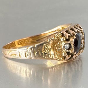 May include: A gold ring featuring a central dark oval gemstone flanked by small white pearls. The band is crafted from yellow gold and displays intricate engraved patterns along the sides, with visible hallmarks stamped on the inner surface.