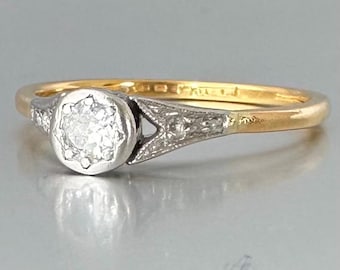 May include: A gold ring featuring a central round white stone set in a silver-toned circular mount. The shoulders of the ring are accented with small white stones embedded in a triangular silver-toned setting that connects to the gold band.