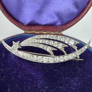 Könnte beinhalten: Silberfarbene Brosche in Form eines Sternschnuppen-Designs, besetzt mit zahlreichen klaren Edelsteinen. Die Brosche hat einen Stern am Anfang des geschwungenen Designs und wird in einer samtgefütterten Schatulle präsentiert.
