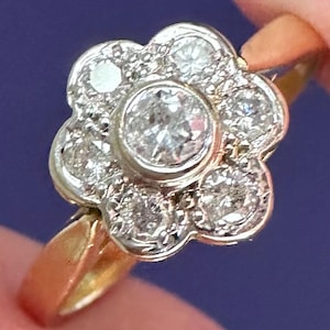 May include: A gold ring featuring a flower-shaped cluster setting made of white metal. The center of the flower is a round, bezel-set clear stone, surrounded by six smaller, round, prong-set clear stones arranged in a circular petal pattern.
