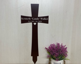 以下が含まれることがあります： ハートの切り抜きと「Kenneth 'Gandy' Walker 19.2.1941 - 18.1.2024」の刻印が施された茶色の金属製の十字架。十字架は明るい木製の表面に置かれ、その前に小さな鉢植えの植物が置かれている。