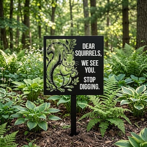 May include: A black metal garden sign featuring a cutout illustration of a squirrel holding an acorn and the white text: DEAR SQUIRRELS, WE SEE YOU. STOP DIGGING. The sign is mounted on a thin black stake positioned in a garden bed.
