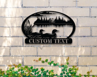 May include: Black metal silhouette of two loons swimming in a lake with a cabin in the background. The silhouette is oval shaped and has the text "CUSTOM TEXT" below it.