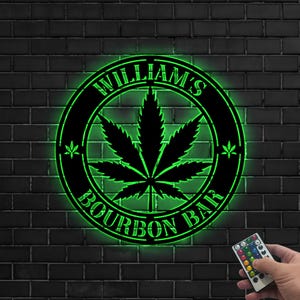 May include: A black metal sign with the words "WILLIAM'S BOURBON BAR" in a circular design, featuring a cannabis leaf in the center. The sign is illuminated with green LED lights and mounted on a dark brick wall.