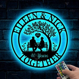 May include: A black metal silhouette of a couple sitting on a bench under a tree with the text "Helen & Nick 40 Years Together" around the outside. The silhouette is illuminated with a blue light.