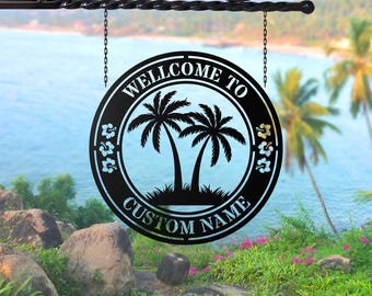 Könnte beinhalten: Schwarzes Metallschild mit den Worten "WELCOME TO" und "CUSTOM NAME", die ein Palmenmotiv umgeben. Das Schild hängt an einer dekorativen Rankenhalterung mit Ketten vor einem tropischen Strandhintergrund.