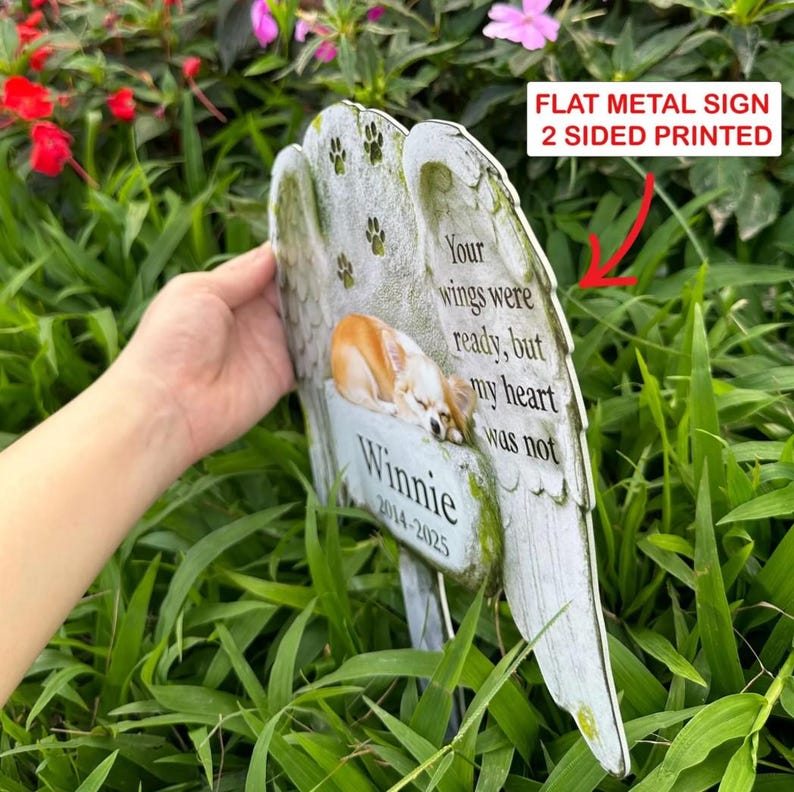 May include: A flat, two-sided metal memorial sign shaped like angel wings. It features a printed image of a tan dog, the text "Your wings were ready, but my heart was not," the name "Winnie," and the years "2014-2025" with paw print accents.