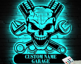 May include: A black and turquoise garage sign featuring a skull with crossed wrenches and pistons. The sign has the text "CUSTOM NAME GARAGE" and is illuminated with turquoise light. A remote control is in the lower right corner.