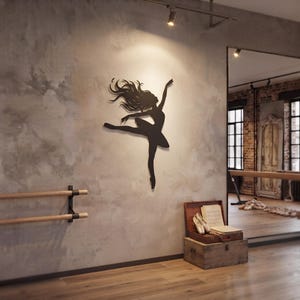 May include: A black metal wall art piece depicting a silhouette of a dancer in a dynamic pose with flowing hair, mounted on a textured gray wall. The figure is captured mid-leap with one arm extended upward and one leg extended outward.