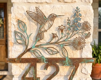 May include: A metal house number sign featuring the digits 427 in a rusted brown finish with teal patina accents. Above the numbers, a decorative cutout depicts a hummingbird hovering near a branch with leaves and various flower blossoms.