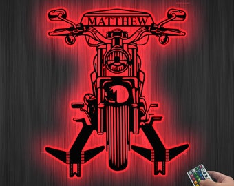 Pode incluir: Uma silhueta vermelha iluminada de uma motocicleta com o nome "MATTHEW" no assento. A motocicleta tem um farol dianteiro e um pneu traseiro. A silhueta está sobre um fundo de madeira escura. Um controle remoto com vários botões coloridos está no canto inferior direito da imagem.