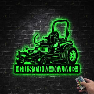 Op de afbeelding: Zwart en groen verlicht metalen bord met een grasmaaier en de tekst "CUSTOM NAME" op een zwarte bakstenen muur. Het bord wordt bediend met een afstandsbediening.
