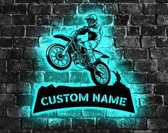 May include: Black metal wall art of a motocross rider on a motorcycle, with the text "CUSTOM NAME" below. The design is illuminated with a turquoise glow against a dark brick wall background.