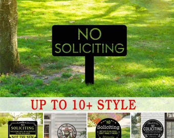 May include: A black metal "No Soliciting" sign with a stake in a grassy garden. The sign has the words "NO SOLICITING" in large, bold letters. Additional sign styles are shown below the main sign.