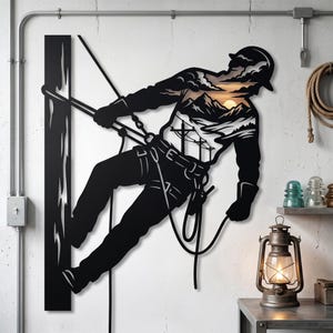 May include: Black metal wall art depicting a lineman working on a power pole. The silhouette of the worker is filled with a landscape scene of mountains, power lines, and a sunset. The art piece is mounted on a white wall.