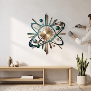 May include: A celestial-themed metal wall art piece featuring a central sunburst design surrounded by orbiting spheres and elliptical rings in shades of metallic gold, teal, and dark green, with sharp geometric spikes radiating outward.