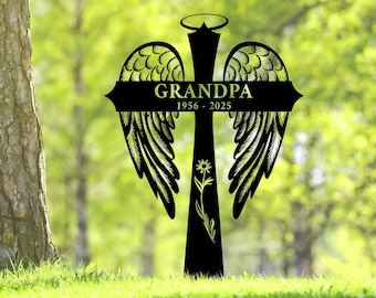 May include: Black metal memorial cross with angel wings, the word "GRANDPA" and the years 1956-2025. A floral design is on the cross. The cross is set in green grass with a blurred green background.