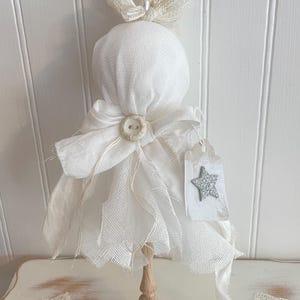 May include: A white fabric ornament featuring a rounded top, a central button, and layered mesh fabric. It is topped with a ribbon loop, a small pearl, and dried foliage. A rectangular tag with a silver glitter star hangs from the side, all mounted on a small wooden spindle base.