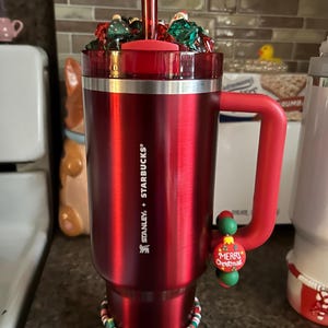 May include: A red stainless steel travel mug with a black lid and a red handle. The mug has a white and green Christmas-themed band around the bottom and a green and red "Merry Christmas" charm attached to the handle. The mug is filled with red and green candy and a red straw.