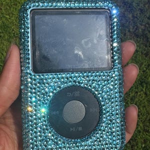 May include: A portable digital music player featuring a rectangular screen and a circular navigation wheel, with the entire outer casing covered in small, light blue, reflective rhinestones that create a sparkling, textured surface appearance.