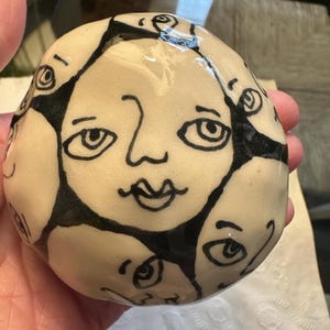 以下が含まれることがあります： 黒いインクで複数の顔が描かれたセラミックの球体。顔は大きな目、鼻、口をしています。球体は薄いベージュ色です。