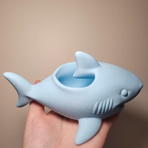May include: A light blue shark-shaped planter. The planter has a round opening on its back and a fin on its top. The shark has a smiling mouth and a small eye on each side. The planter is held in a hand.