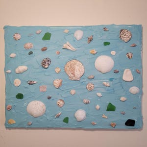 May include: A rectangular artwork featuring a textured, light blue background adorned with various seashells, sea glass, and small stones. The seashells vary in size, shape, and color, creating a beach-themed composition. The artwork is likely a decorative piece.