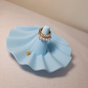Könnte beinhalten: Ein hellblauer Keramik-Ringständer in Form einer Muschel mit drei goldenen Ringen mit Perlenbesatz.
