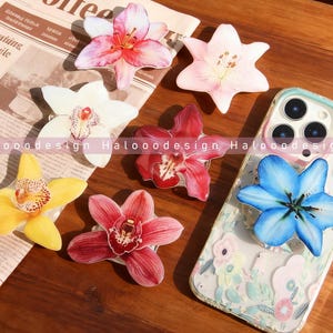 Peut inclure: Plusieurs supports de téléphone en forme de fleurs, aux couleurs rose, rouge, jaune, blanc et bleu. Les supports ressemblent à des orchidées et des lys. L'un d'eux est fixé à un téléphone avec une coque florale. Le mot "Coffee" est visible sur un journal.