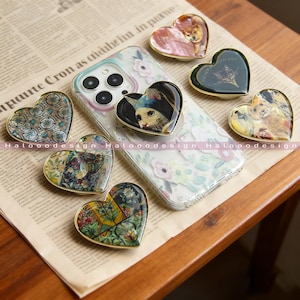 May include: Heart-shaped phone grips with diverse designs, such as floral patterns and animal portraits, including a cat wearing a hat. The grips have gold edges and are arranged on a newspaper.