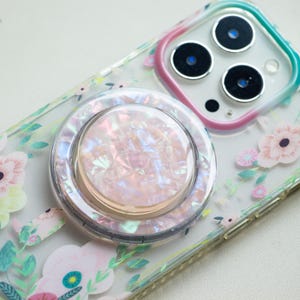 May include: A clear phone case with a floral pattern and a pearlescent phone grip. The phone grip is circular with a pink and white iridescent finish. The case has a pink, green, and blue border around the camera.