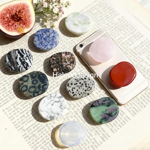 Könnte beinhalten: Eine Sammlung verschiedener Handyhalterungen aus Edelsteinen in verschiedenen Farben und Mustern, darunter Blau, Weiß, Schwarz und Rot. Ein Smartphone mit einem rosa Halter ist ebenfalls sichtbar. Das Bild ist auf einer Vintage-Buchseite platziert.