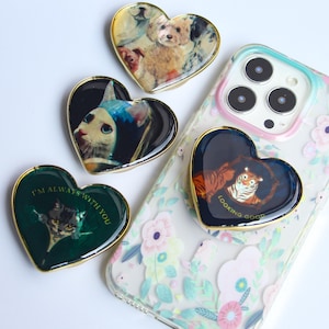 May include: Heart-shaped phone grips with gold edging, each displaying an animal portrait. One grip has a tiger with the text "LOOKING GOOD". Another has a cat with the text "I'M ALWAYS WITH YOU". The grips are on a floral phone case.