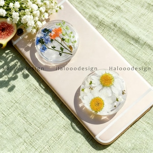 May include: Two clear, round phone grips with embedded dried flowers. One grip features blue and orange flowers, the other has white daisies with yellow centres. The grips are on a light-coloured phone.