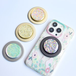Op de afbeelding: Een verzameling glitter telefoonhouders in goud, zilver en zwart. Eén houder is bevestigd aan een telefoonhoesje met bloemenpatroon. De houders hebben kleurrijke glitterontwerpen in verschillende tinten.