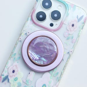 May include: A clear phone case with a floral pattern and a light purple phone ring holder. The ring holder has a pearlescent centre and a rose gold trim. The phone case is decorated with pink, yellow, and white flowers.