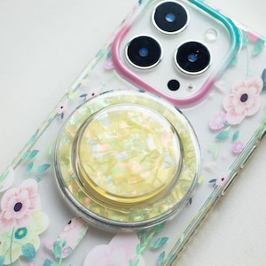 May include: A clear phone case with a floral design and a pearly white phone ring holder. The case has a pink and green accent around the camera lens.