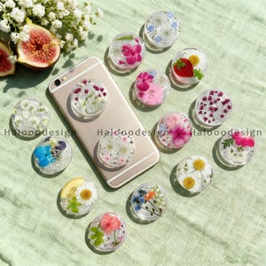 Può includere: Una collezione di supporti per telefono rotondi e trasparenti con vari fiori secchi e frutta. I supporti sono disposti attorno a uno smartphone dorato su una superficie verde chiaro. Alcuni supporti presentano margherite, fragole e altre composizioni floreali colorate.