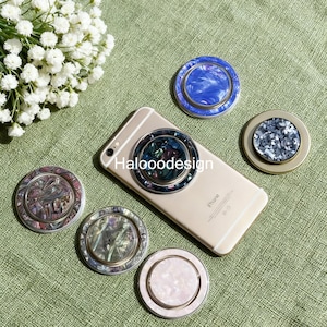May include: A selection of decorative phone ring holders with iridescent and marbled patterns. A phone has a ring holder attached. The ring holders are in shades of blue, pink, and grey, with gold and silver accents. The text "Halooodesign" is visible.