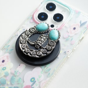 以下が含まれることがあります： ターコイズとシルバーの電話グリップ。蹄鉄のデザインとカウボーイブーツのペアが特徴です。グリップは、花柄のクリアな電話ケースに取り付けられています。