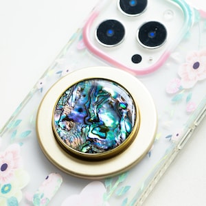 May include: A gold phone ring holder with a iridescent abalone shell inlay. The ring holder is attached to a clear phone case with a floral design.