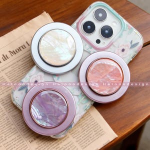 May include: Three circular phone grips with shimmering centres in white, pink, and purple, each with a metallic edge. The grips are on a floral phone case, resting on a newspaper.