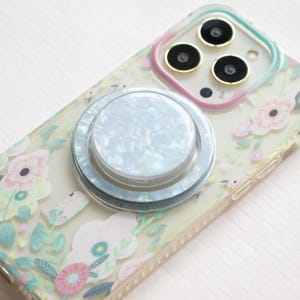 Puede incluir: Una funda transparente para teléfono con un patrón floral y un soporte para anillo de teléfono blanco iridiscente. La funda del teléfono tiene un borde dorado alrededor de las lentes de la cámara.